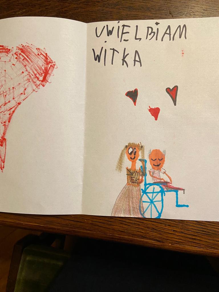 Na zdjęciu widzimy dziecięcy rysukenk z podpisem UWIELBIAM WITKA, serca i dziewczynkę z dziadkiem siedzącym na wózku - oboje się uśmiechają