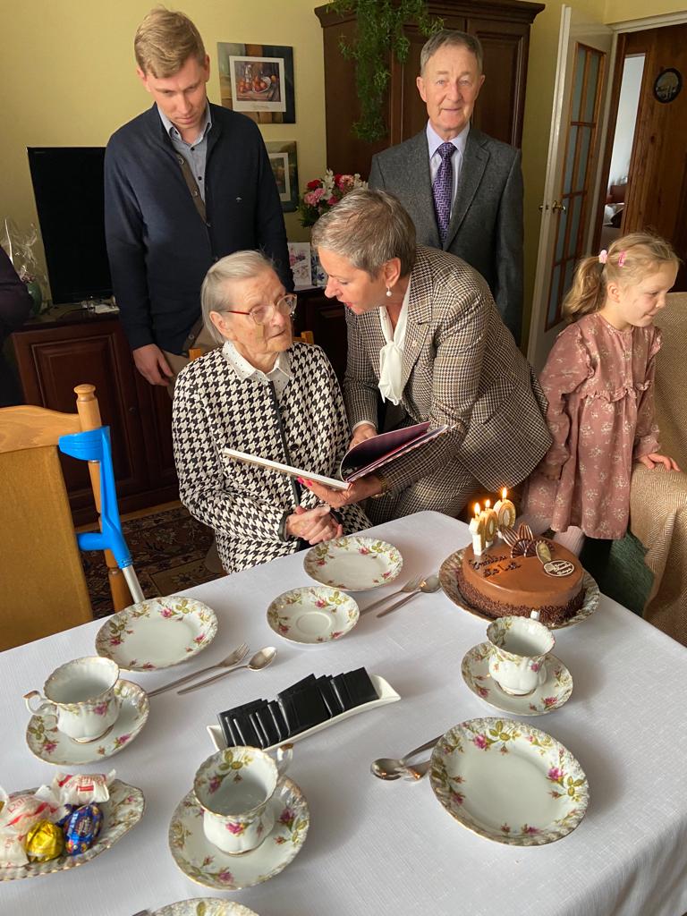 Na zdjęciu widizmy 5 osób - Jubilatkę i Panią Prezydent trzymającą Album o Słupsku, za nimi 2 mężczyzn oraz po prawej stronie dziewczynka. Na pierwszym planie widzimy stół z porcelanowymi filiżankami i talerzykami oraz słodyczami