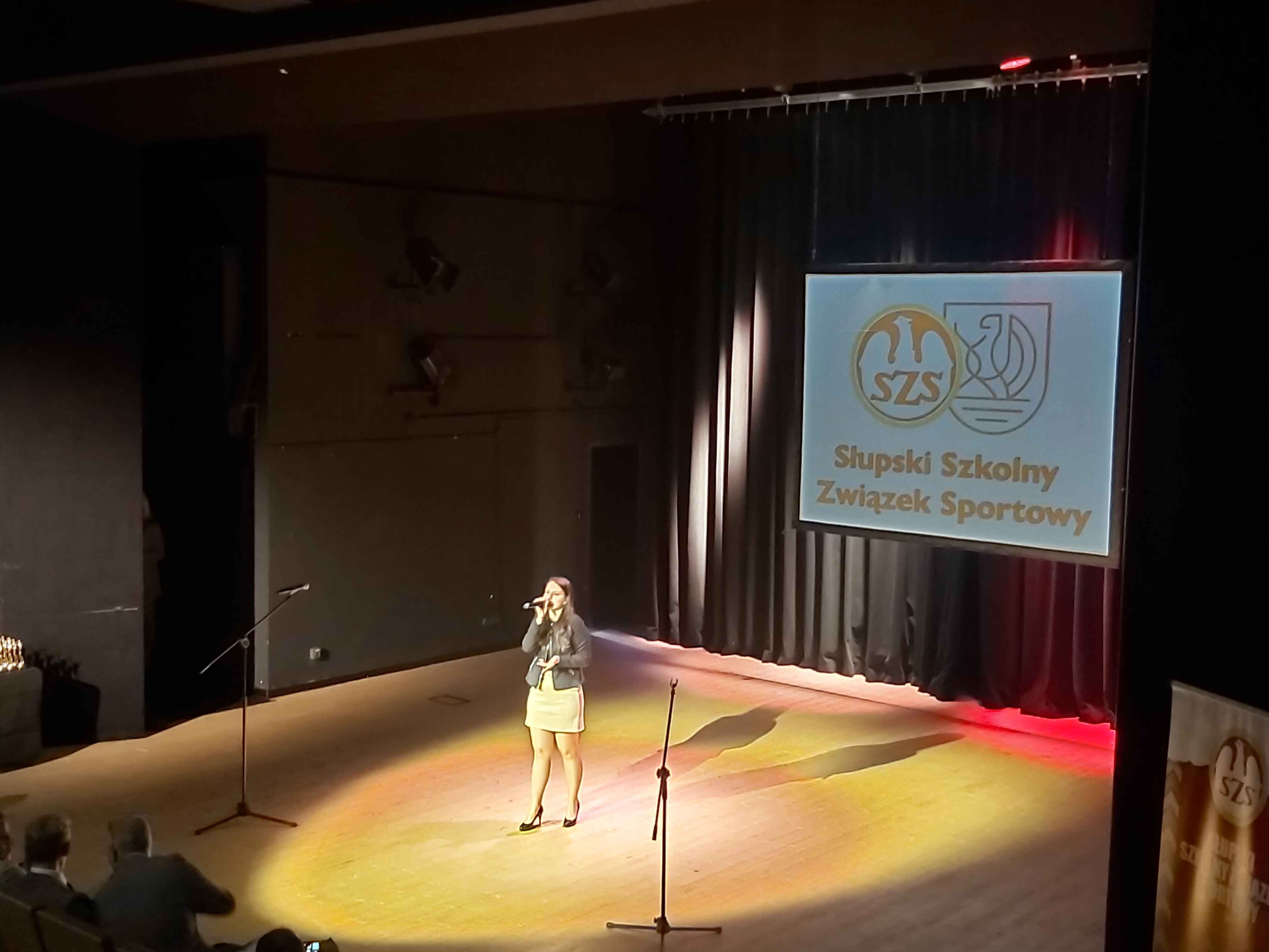 Dziewczyna stojąca na scenie i śpiewająca, w tle napis wyświetlony Słupski Szkolny Związek Sportowy i loga Związku oraz Miasta Słupska