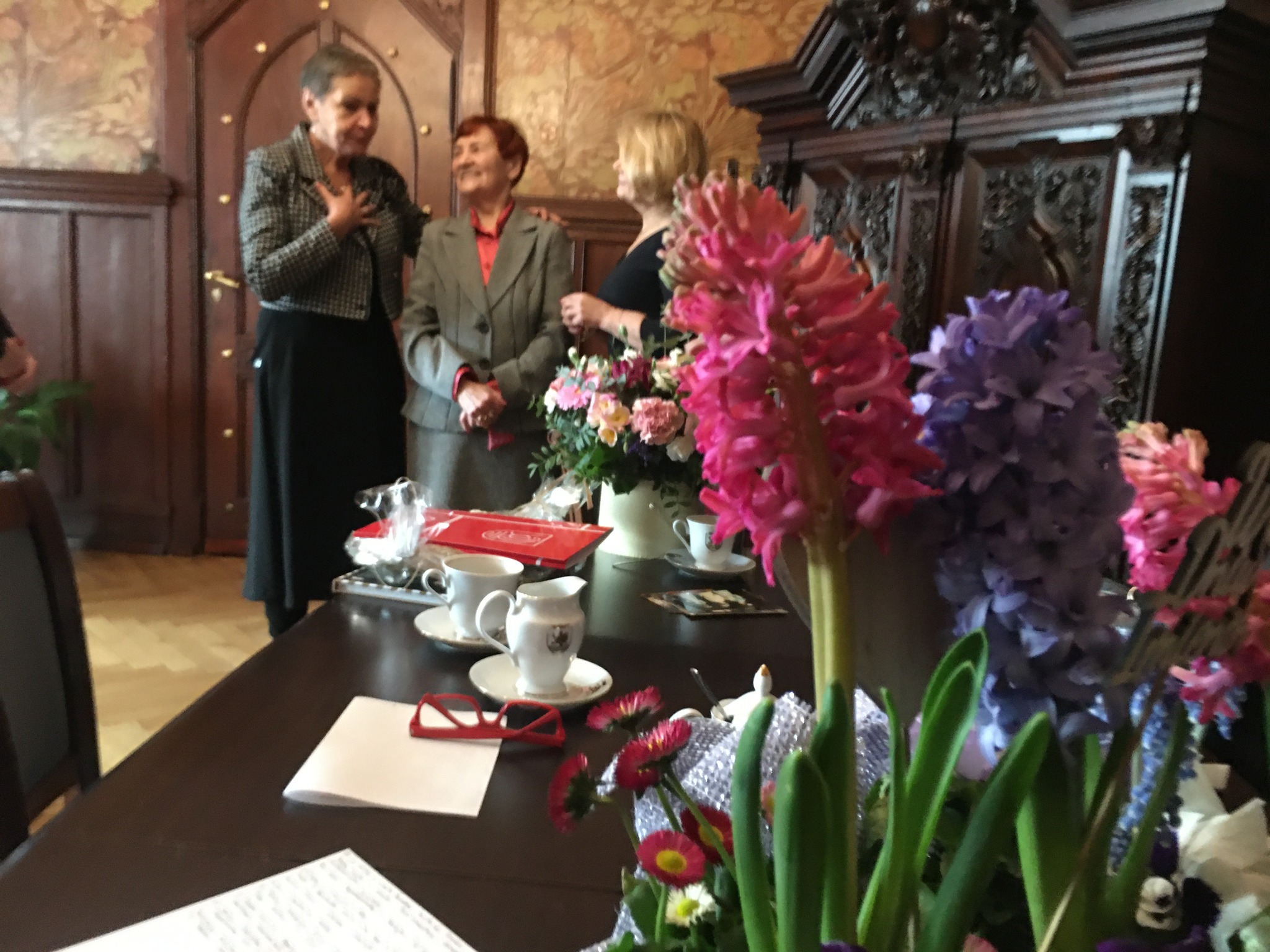 Na pierwszym planie kwiaty, w tle stół, filiżanki i Prezydentka z Krystyną Homendą oraz Violetta Karwalska