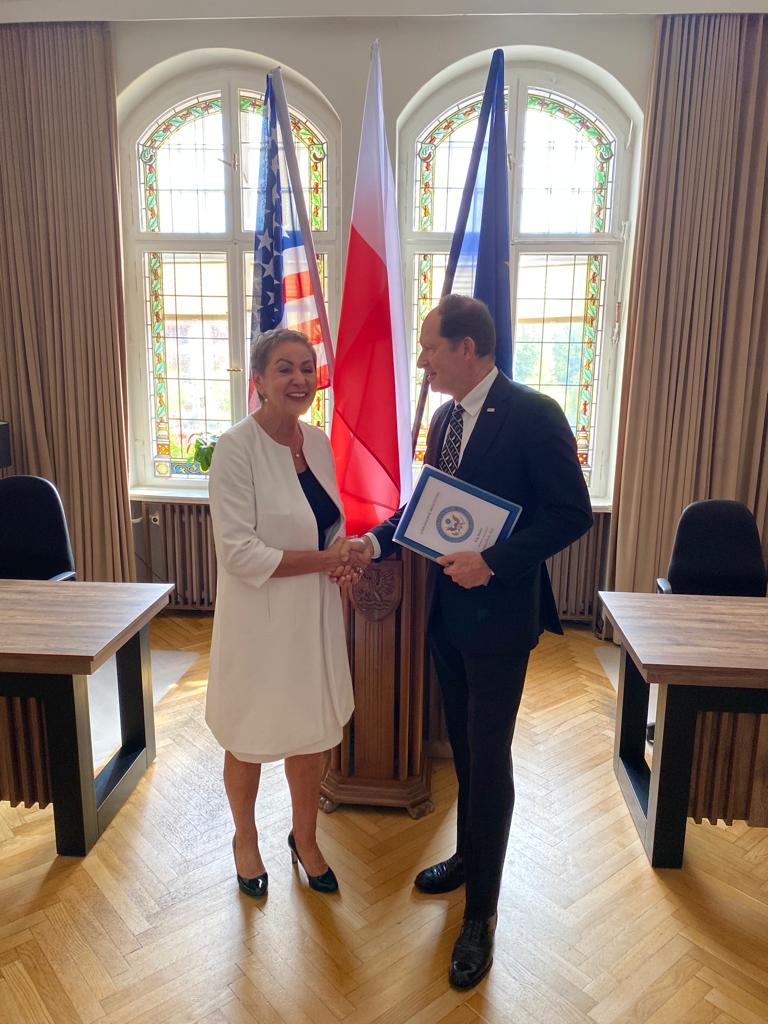 Na zdjęciu widzimy Prezydentkę Miasta i Ambasadora Stanów Zjednoczonych w Polsce. W tle flagi Polski, USA i UE, okna biurka i fotele biurowe