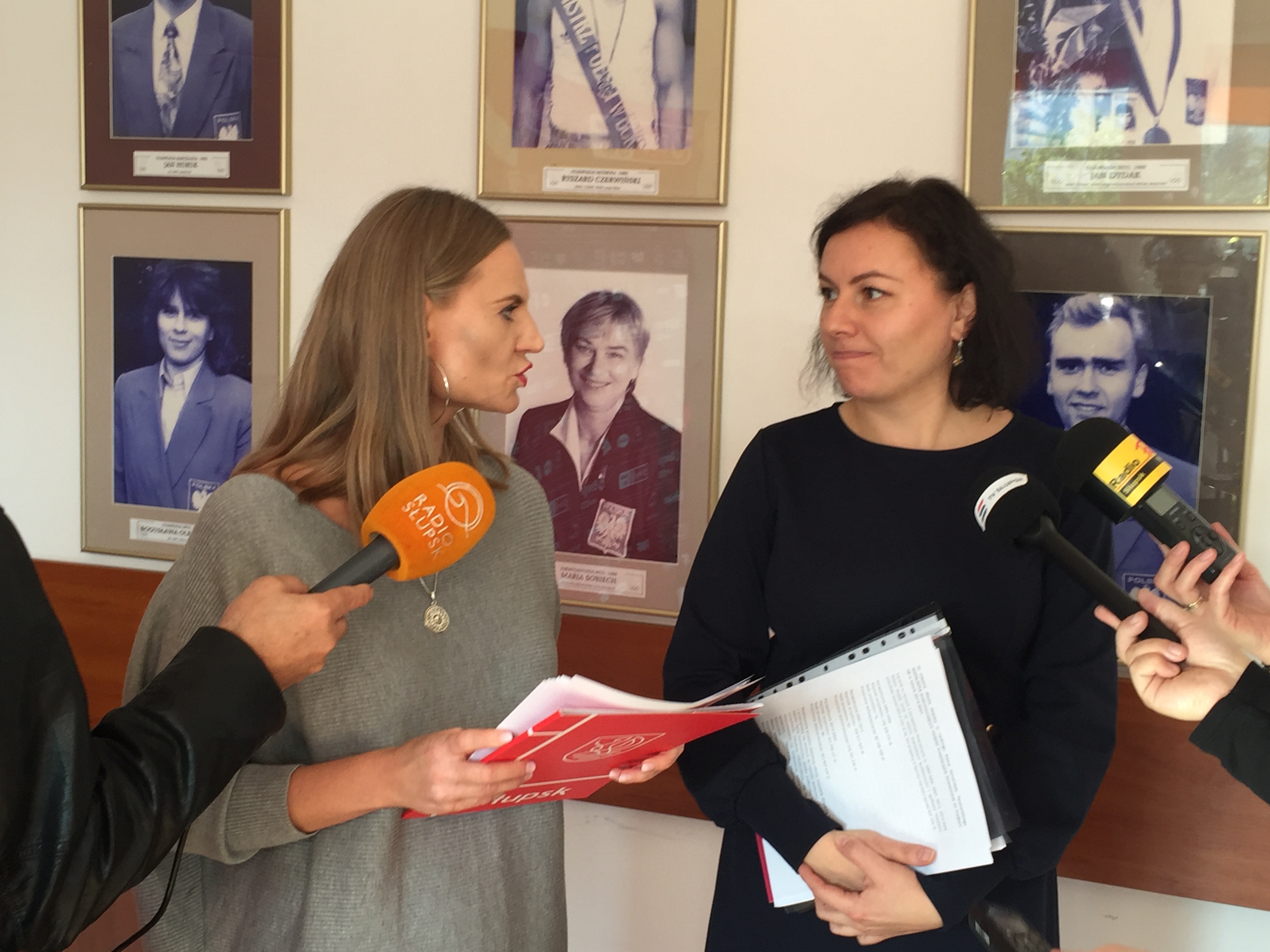 Marta MAKUHC trzyma teczkę obok niej z dokumentami Justyna PLUTA trzymająca dokumenty. Za Paniami widać zdjęcia słupskich olimpijczyków