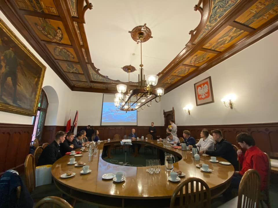 Na zdjęciu widzimy okrągły stół wokół którego siedzą Laureaci i Uczestnicy Festiwalu Explory oraz Prezydenkta miasta. Na ekranie prezenowane jest wideo , w tle widać flagi