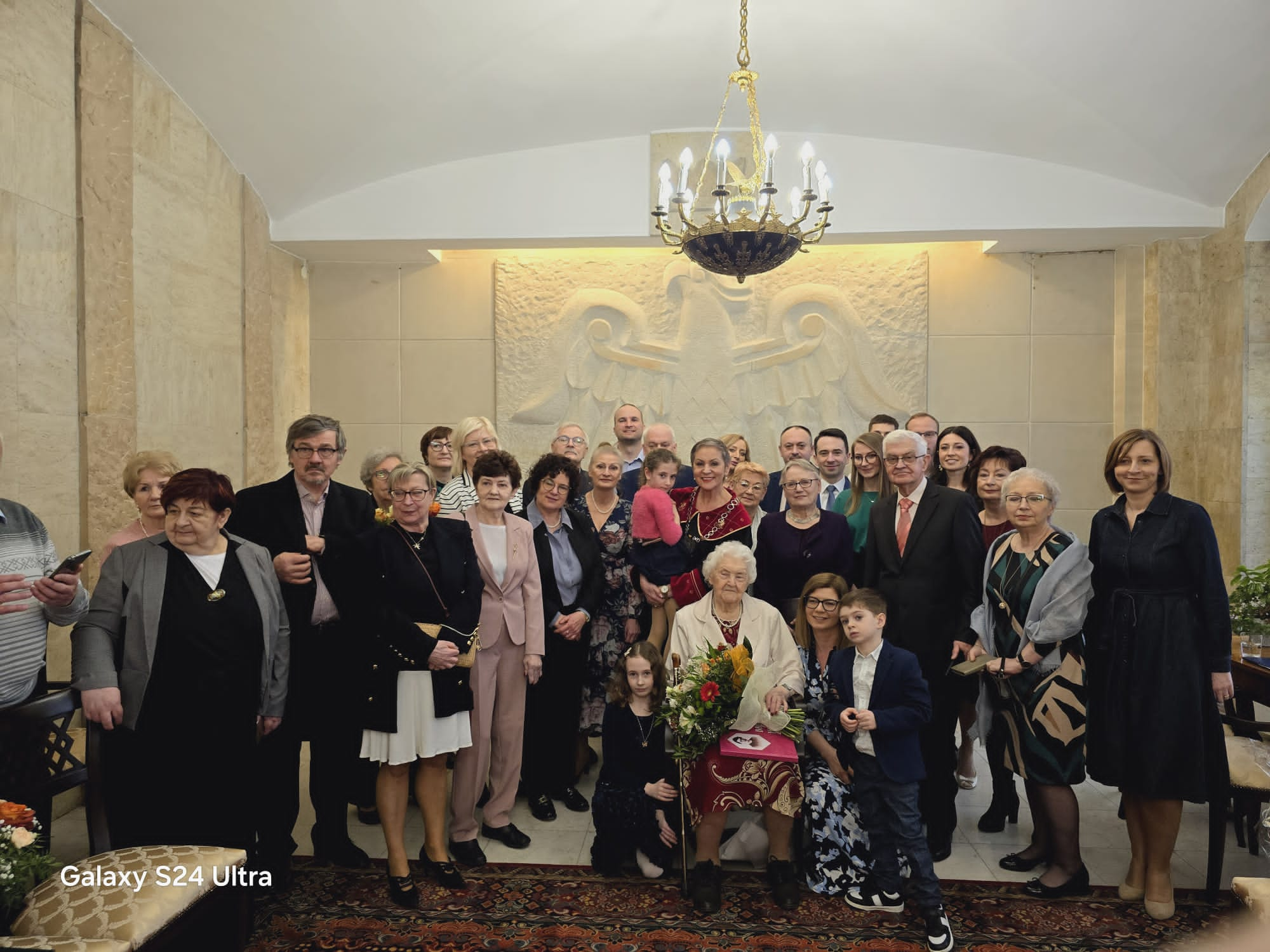 Kilkadziesiąt osób, rodzina i przyjaciele Jubilatki, Pani Prezydent - w Pałacu Ślubów; Jubilatka otoczona bliskimi siedzi na krześle i trzyma upominki