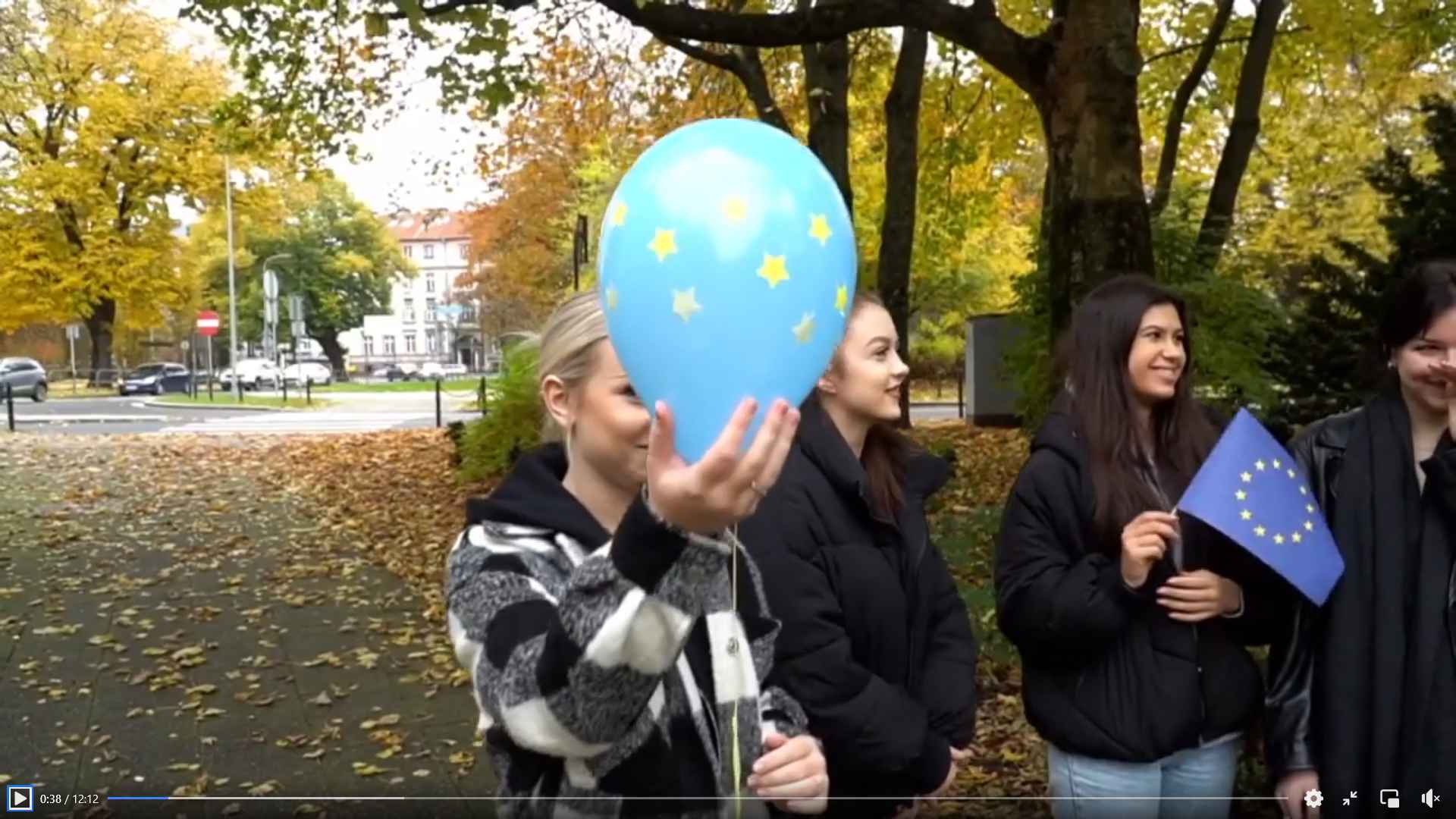 Na zdjęciu wdzimy kilka osób w parku, w tle przejście dla pieszych chodnik i liście. Jedna z dziewczyn na głównym planie trzyma balon symbolizujący Unię Europejską (niebieski z żółtymi gwiazdkami), a druga dziewczyna trzyma flagę UE