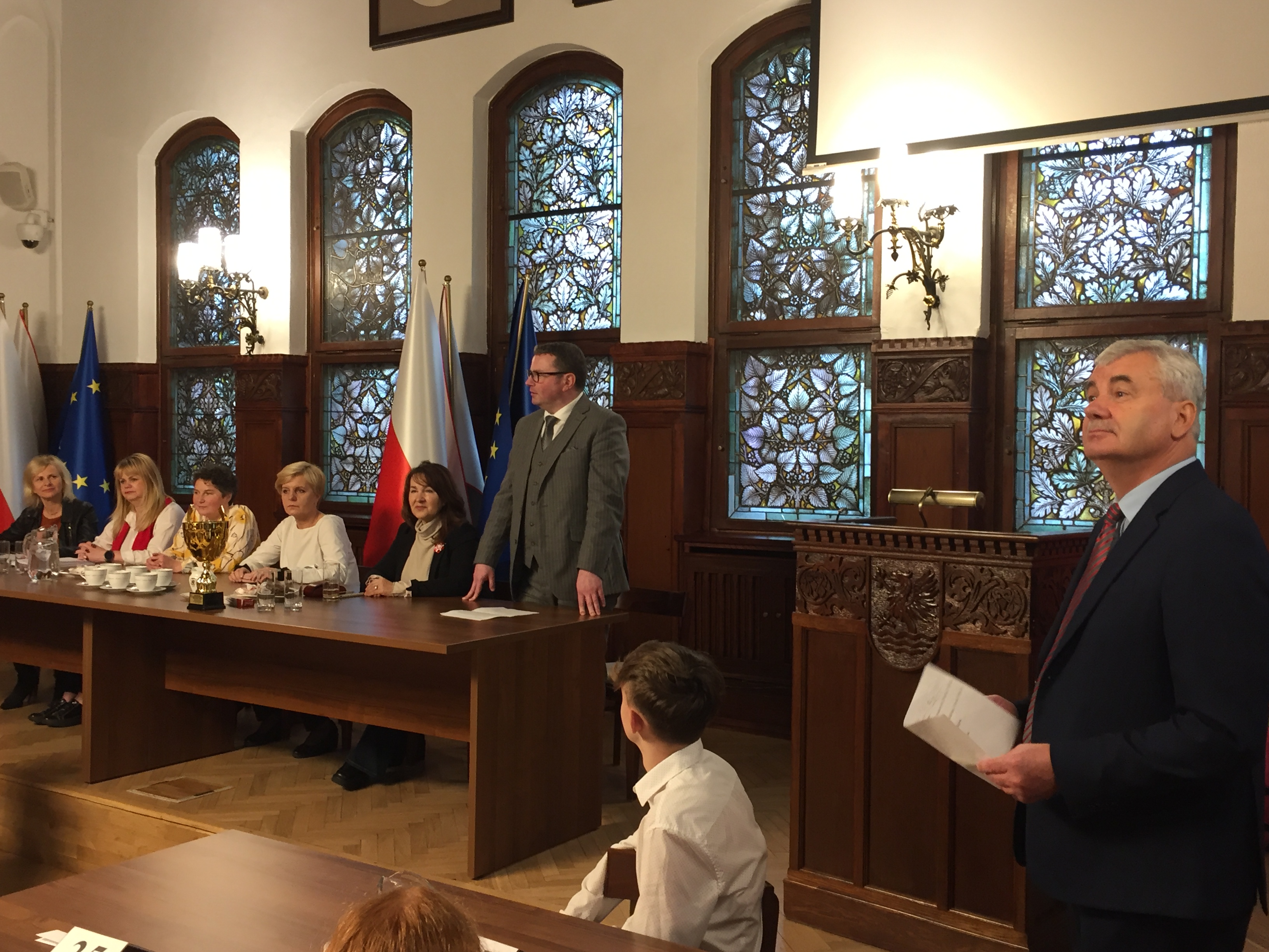Wiceprezydent Golińśki wraz z komisją konkursową oraz Dyrektor SP 10 trzymający kartkę. Oprócz tego stoły, mównica, w tle okna z witrażami