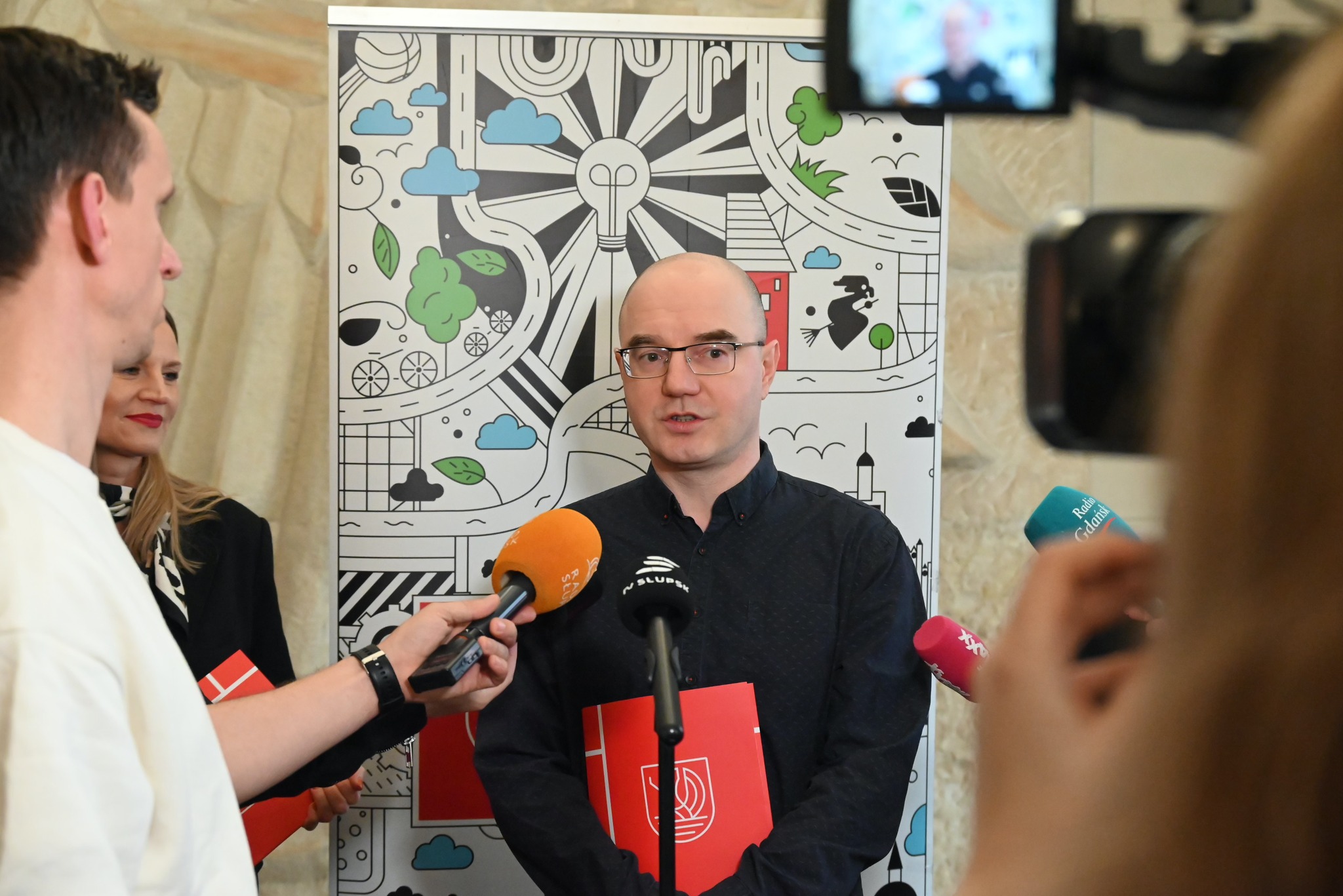 Mężczyzna stoi  przed nim mikrofony przedstawicieli mediów