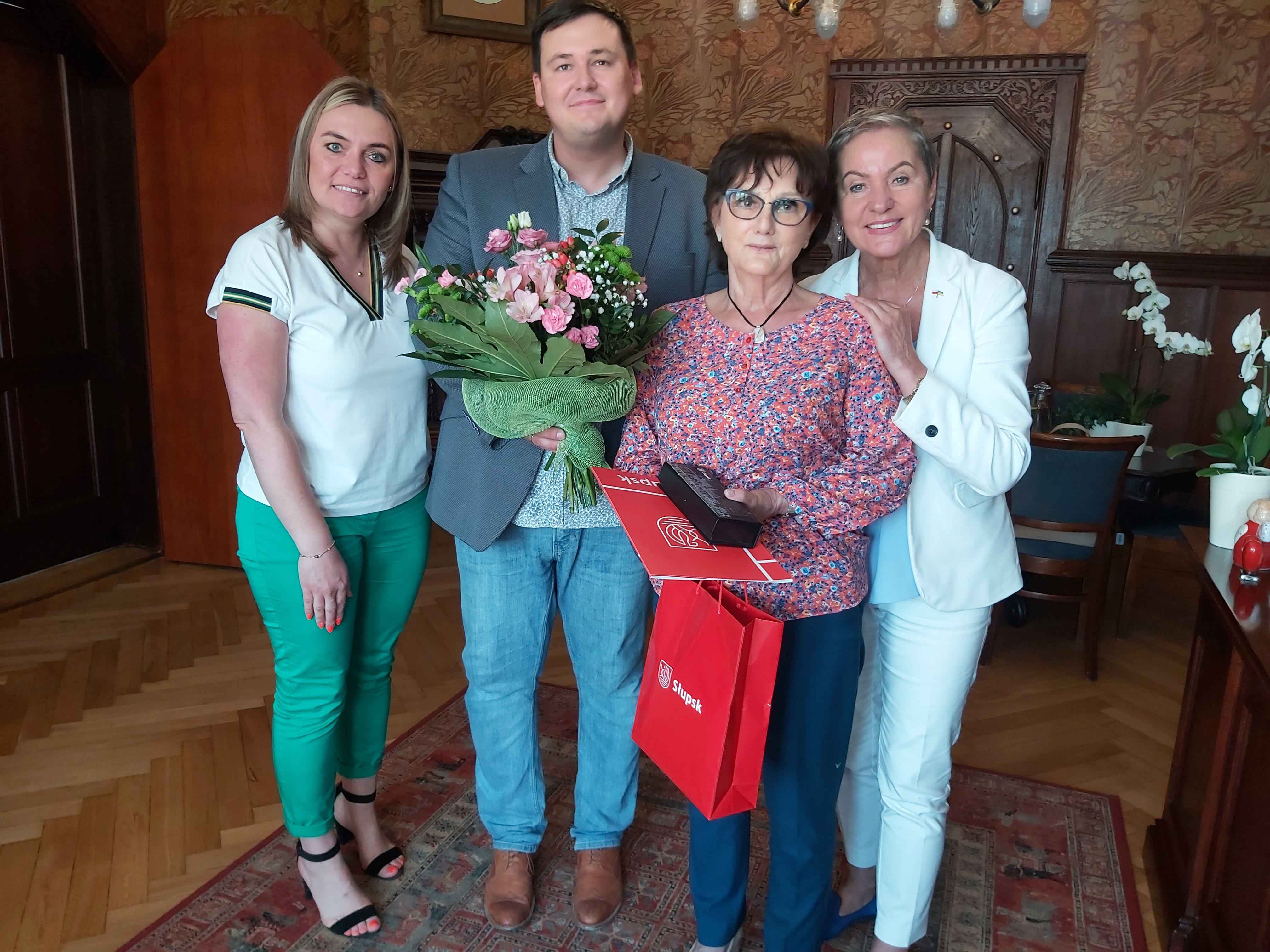 Na zdjęciu widzimy Krystynę Wrycz, wieloletnią Kierowniczkę Urzędu Stanu Cywilnego, trzymającą bukiet kwiatów, teczkę i torbę z upominkiem, a także Panią Prezydent, Panią SKarbnik i Pana Sekretarza