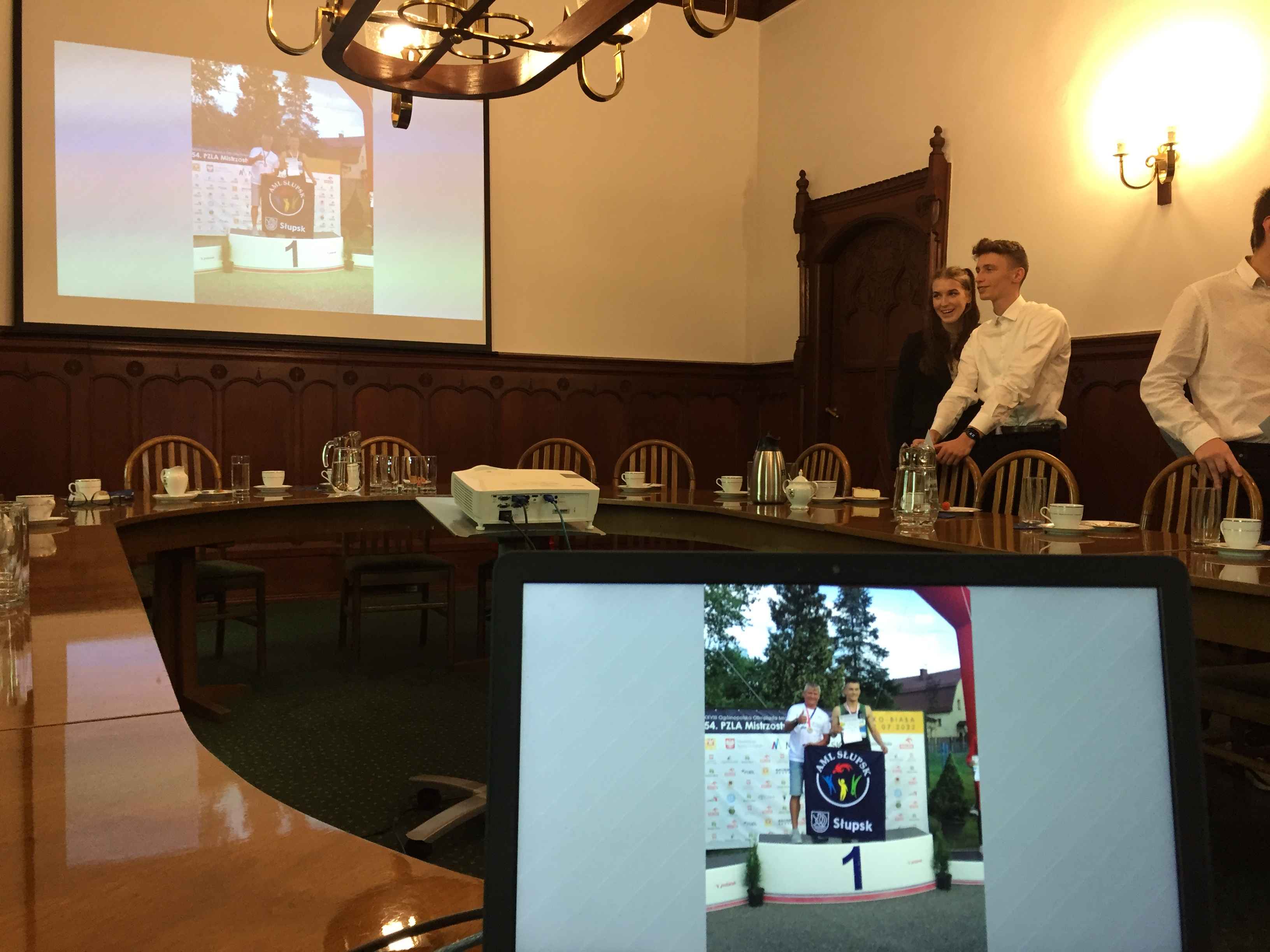 Laptop na pierwszym tle, w głębi ekran wyświetlający zdjęcia słupskich sportowców - teraz wyświetla konkretny slajd biegacz AML z  Trenerem stoją na pierwszym stopniu podium
