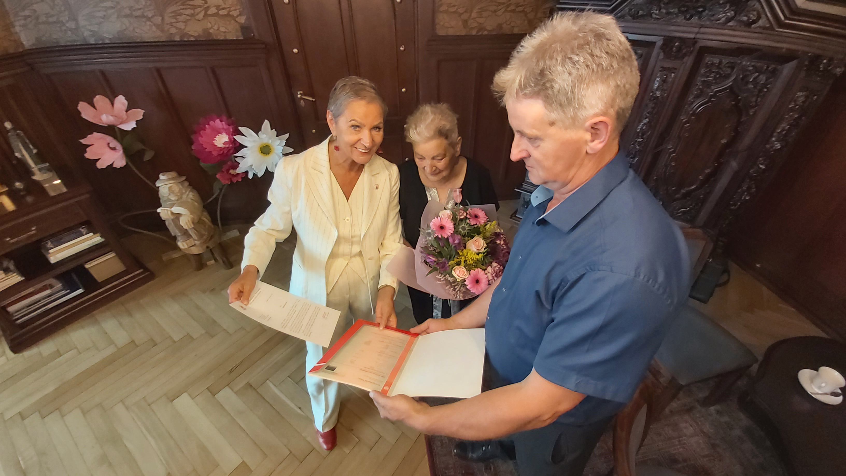 Pani Prezydent wręcza odpis aktu małżeństwa i list gratulacyjny oraz kwiaty. Odbierać upominki Jubilatce pomaga osoba bliska Jubilatki