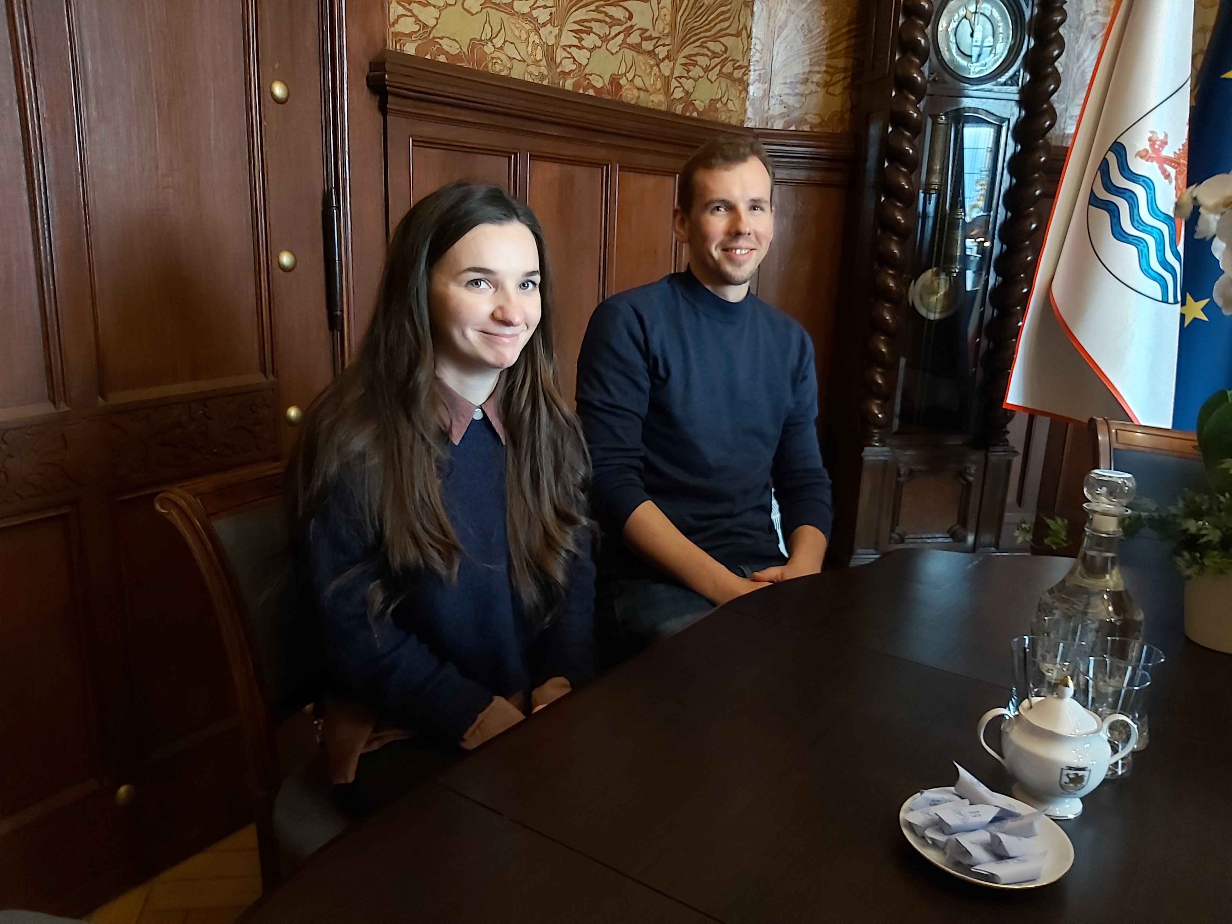 Mistrzowie -Pani Magda i Pan Kamil uśmiechają się, siedząc przy stole, w tle flaga Miasta Słupska oraz zegar