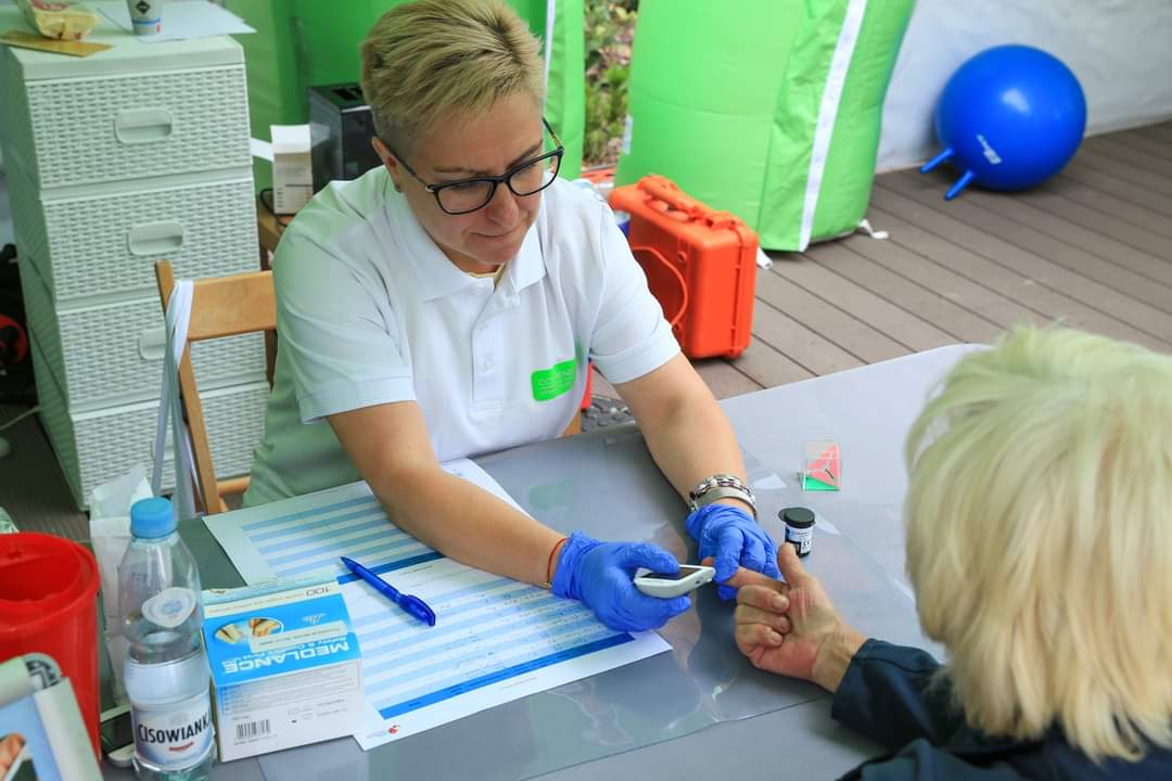 Na zdjęciu widzimy pielęgniarkę wykonującą badania profilaktyczne pacjentce, na stole m.in. woda, kartka, długopis, , fiolka, w tle balon, szafka. Fot. Biblioteka Słupska