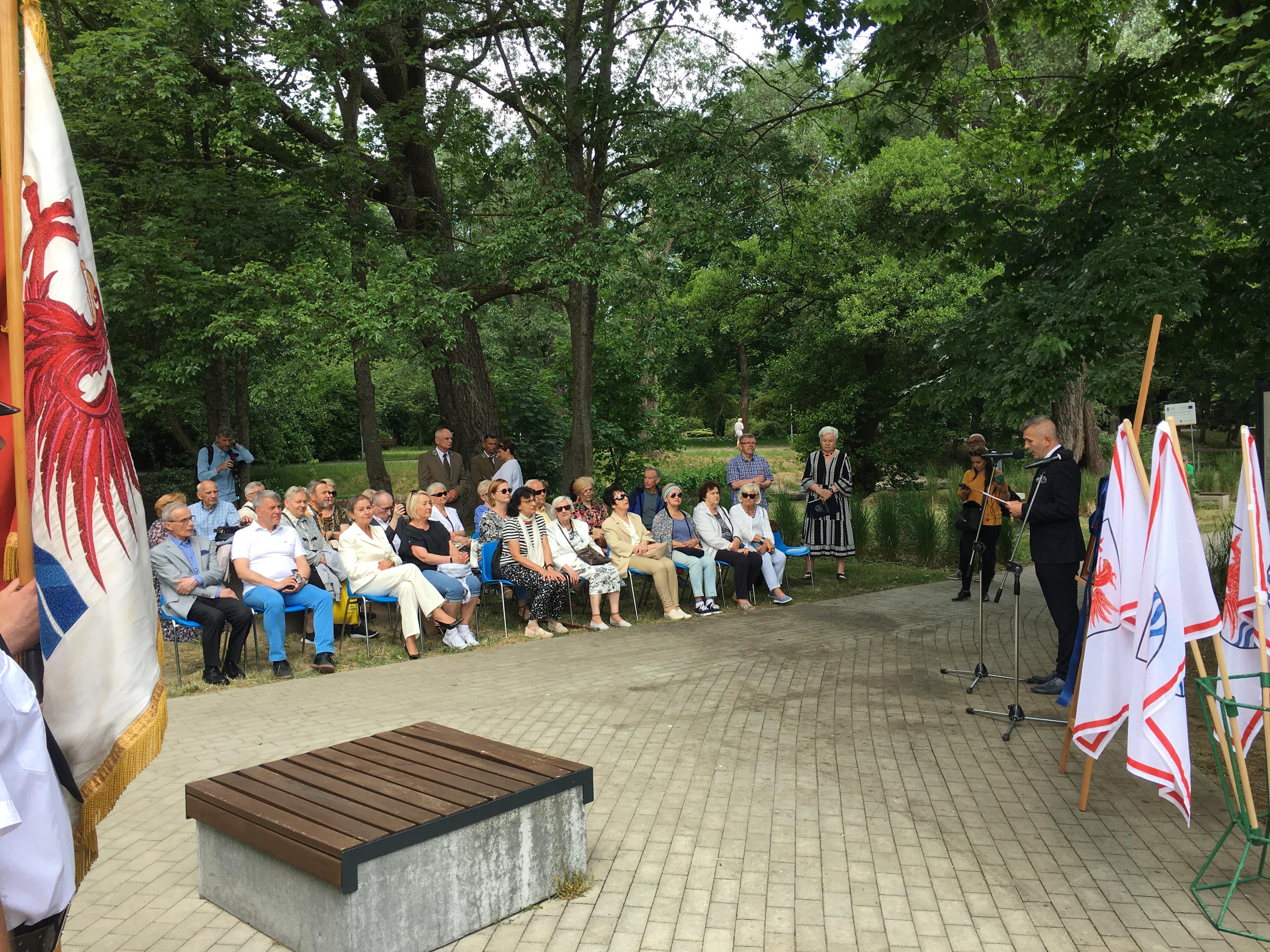 Widok na gości zebranych na uroczystości, prowadzącego uroczystość, Starżników Miejskich oraz flagi miasta Słupska; w tle drzewa i park
