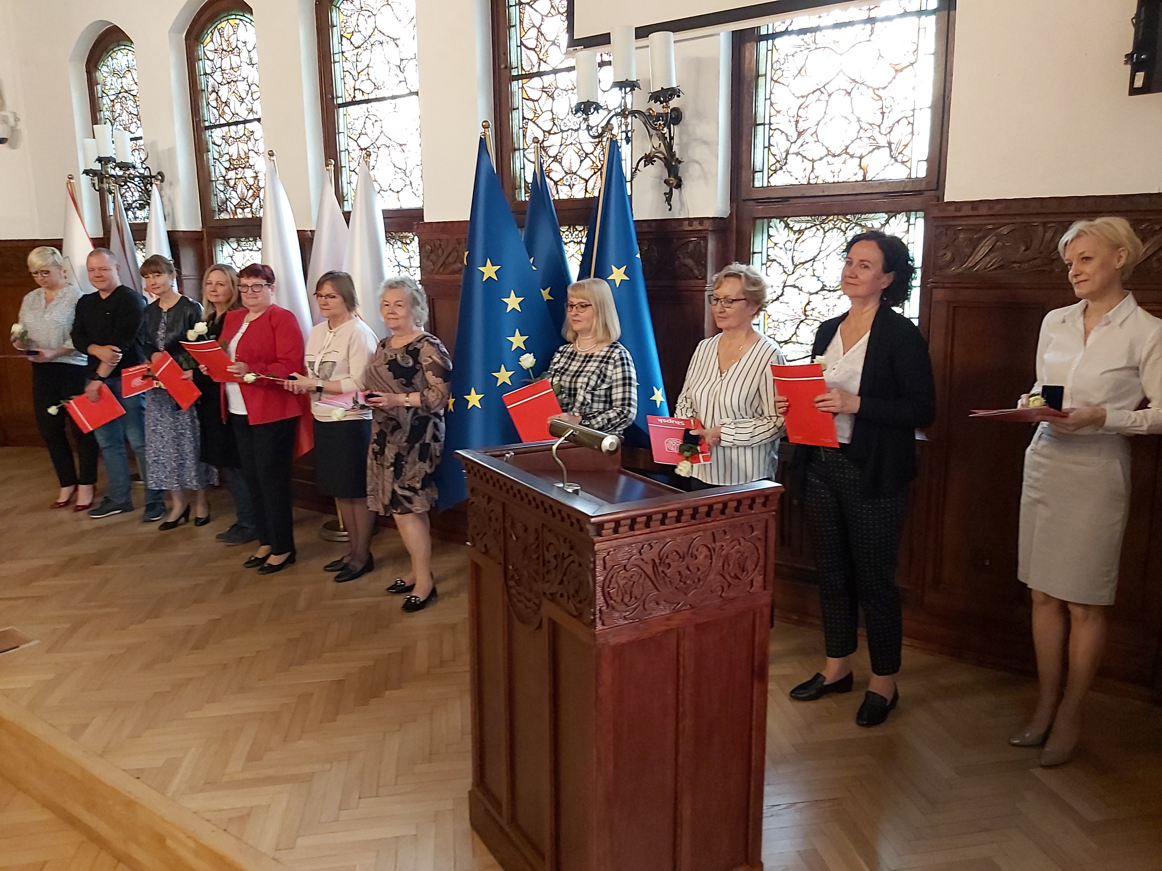 Na zdjęciu widzimy kilkunastu wyróżnionych urzędników, mównicę i  flagi Polski oraz Unii Europejskiej