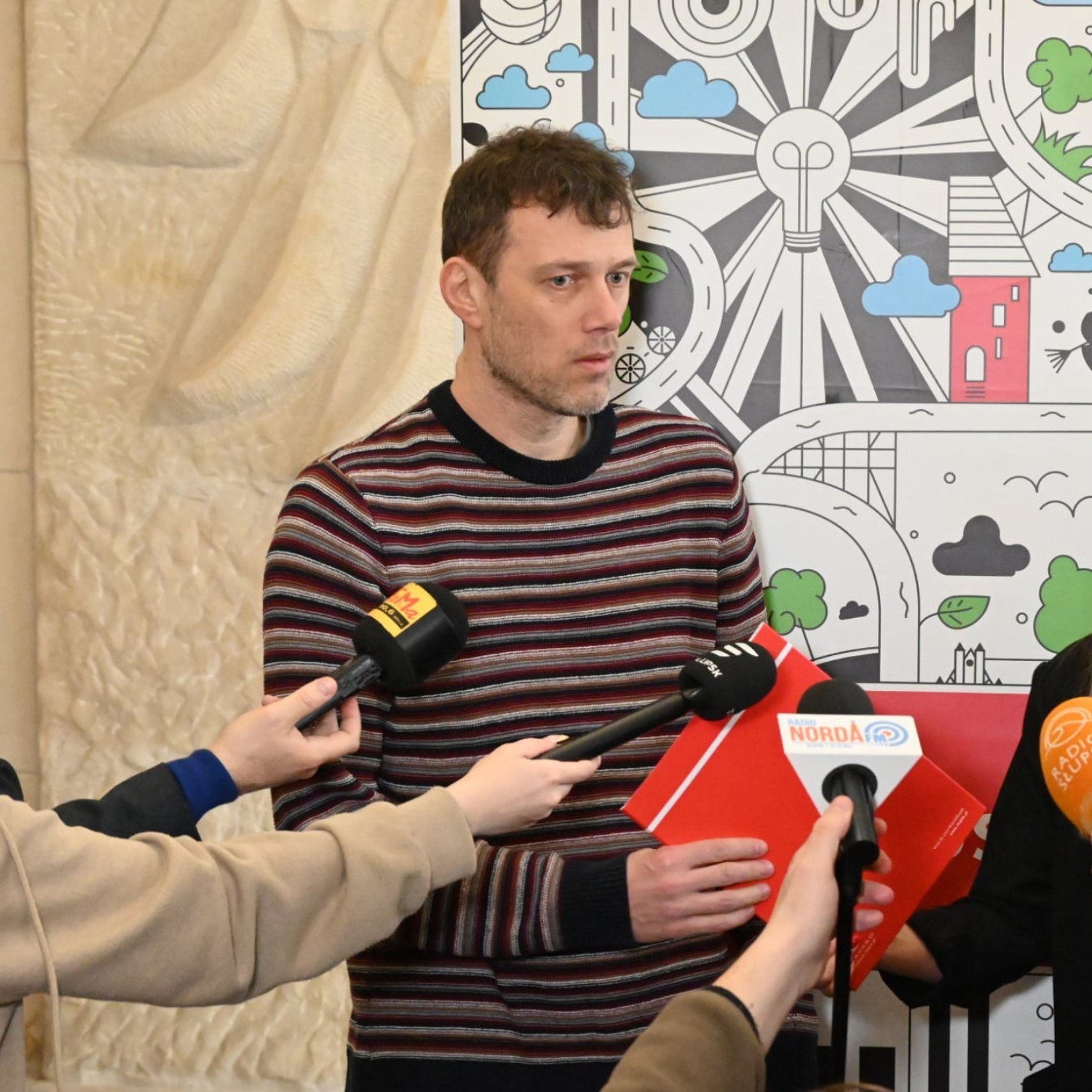 Mężczyzna stoi przed nim mikrofony przedstawicieli mediów trzyma w dłoni czerwony dyplom