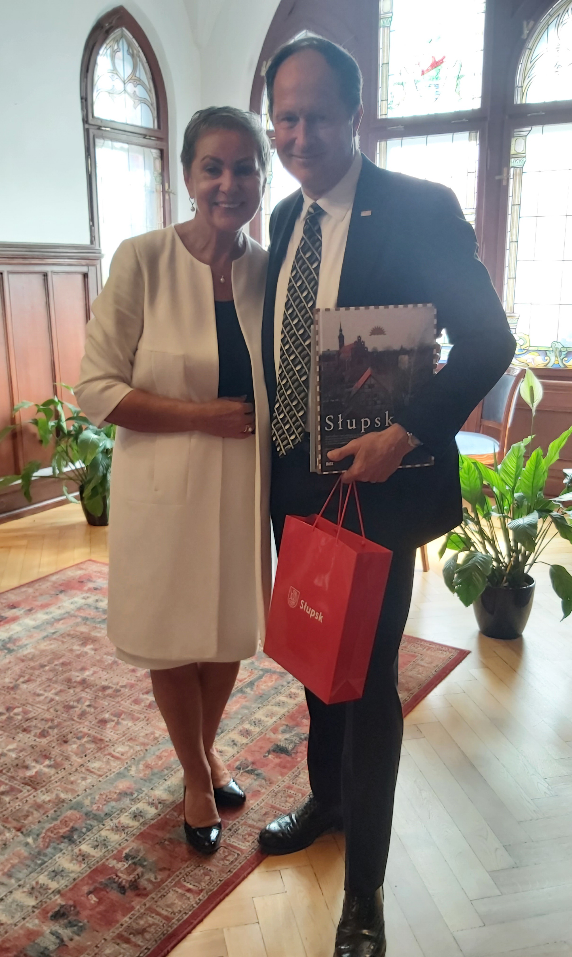 Na zdjęciu widzimy Prezydentkę Miasta i Ambasadora Stanów Zjednoczonych w Polsce trzymającego torbę z prezentami oraz Album o Słupsku, w tle okna i kwiaty w doniczce