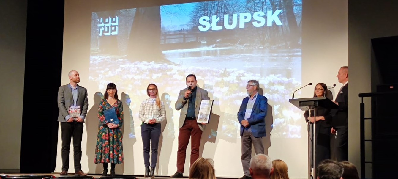 Na zdjęciu widzimy 2 kobiety i 2 mężczyzn odbierających na scenie nagrodę dla Słupska, poza tym 2 innych mężczyzn i kobieta , w tle słupskie kliny zieleni
