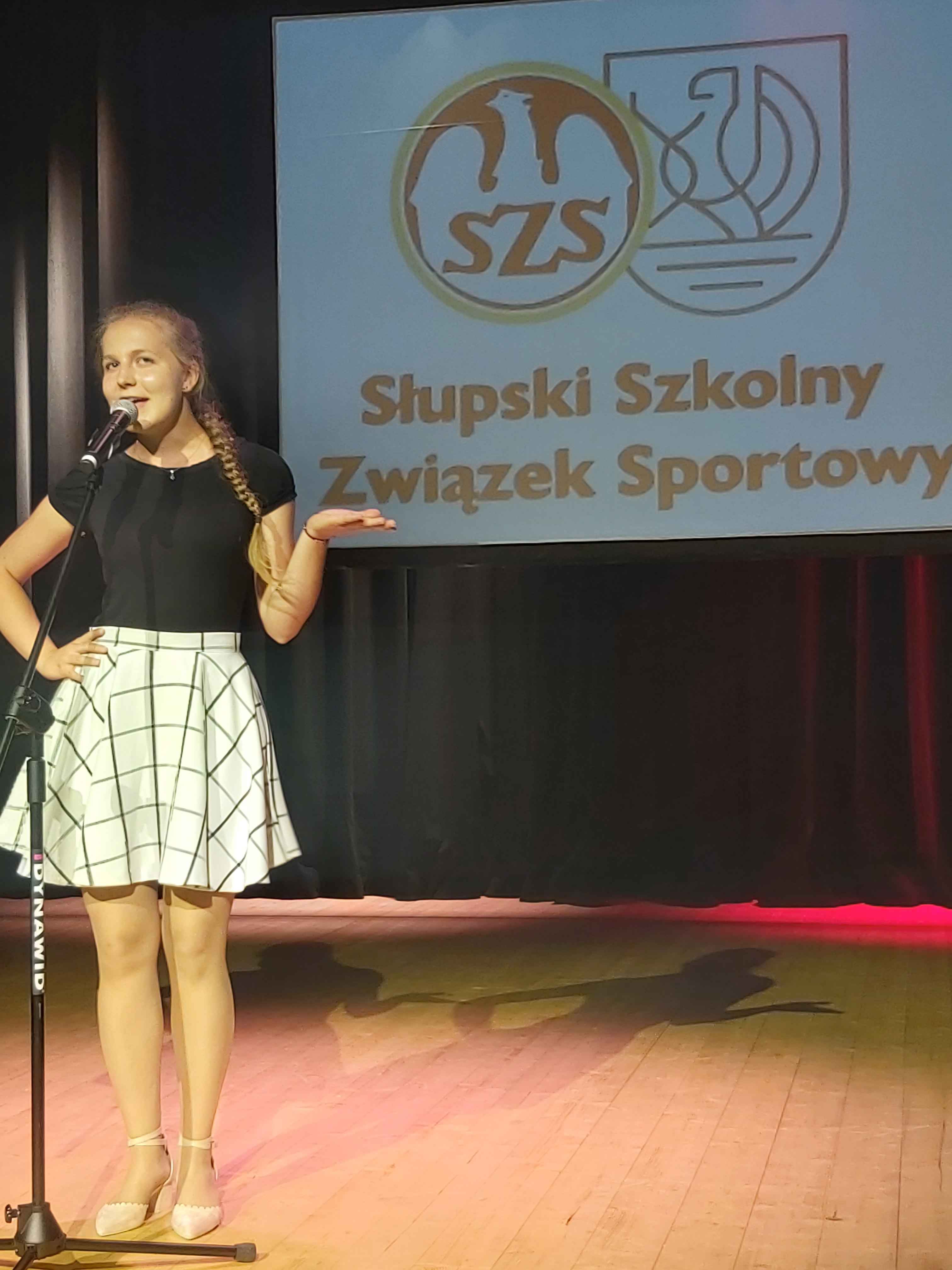 Dziewczyna śpiewa piosenkę stojąc na scenie , w tle wyświetlony napis  Słupski Szkolny Związek Sportowy, na podłodze cień artystki
