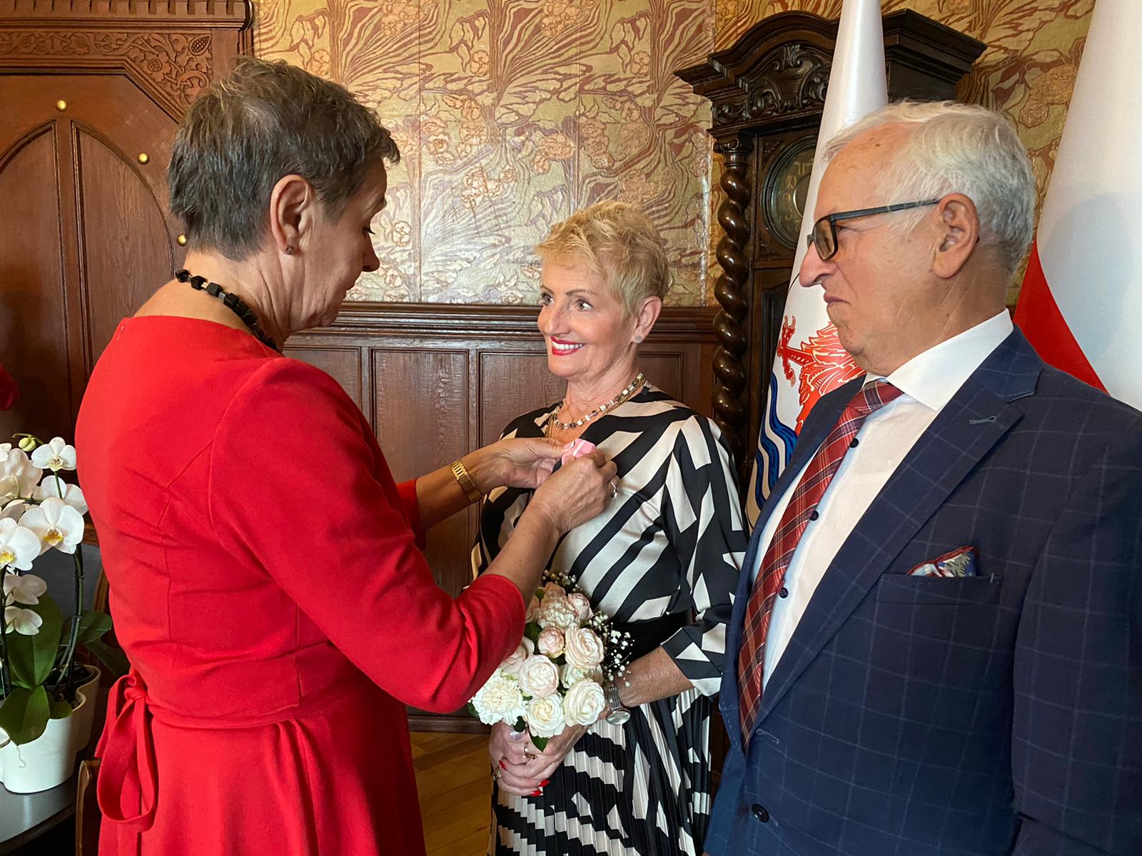 Prezydentka Miasta przypina Jubilatce medal. Obok Jubilat, w tle stół, na którym stoi storczyk, oprócz tego w oddali widać stary zegar i fragment flag