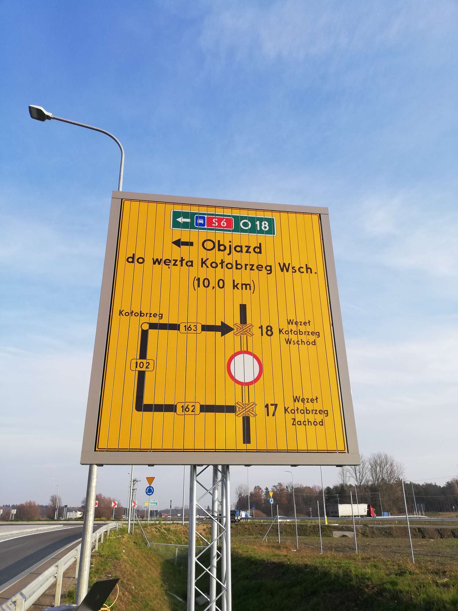 Przykładowa tablica wskazująca objazdy, w tle jezdnia, rondo, znaki drogowe, zieleń, drzewa