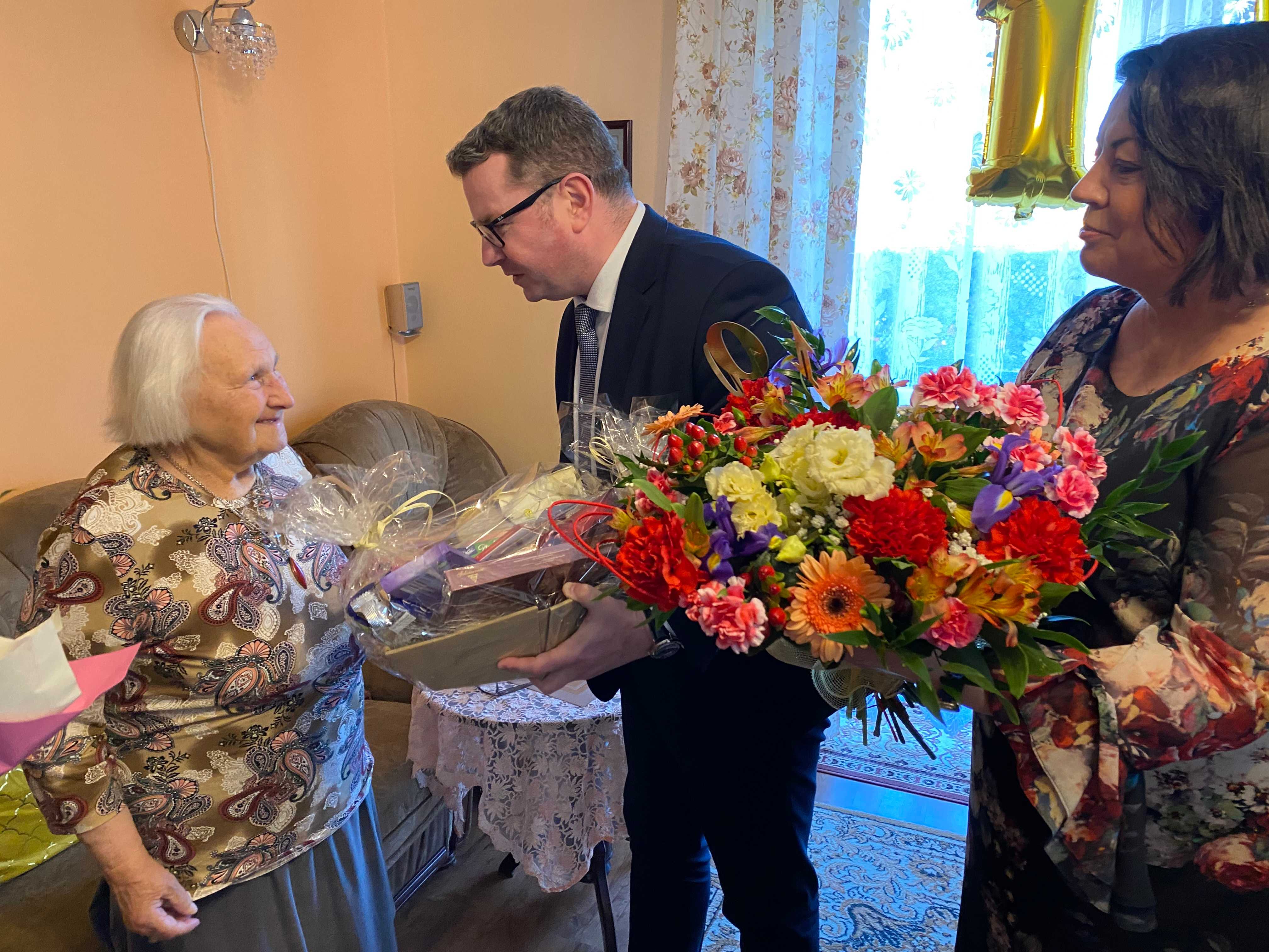 Wiceprezydent Golińśki wręcza Jubilatce kosz z upominkami, obok Pani Kierownik USC trzyma kwiaty, które wręczy Jubilatce