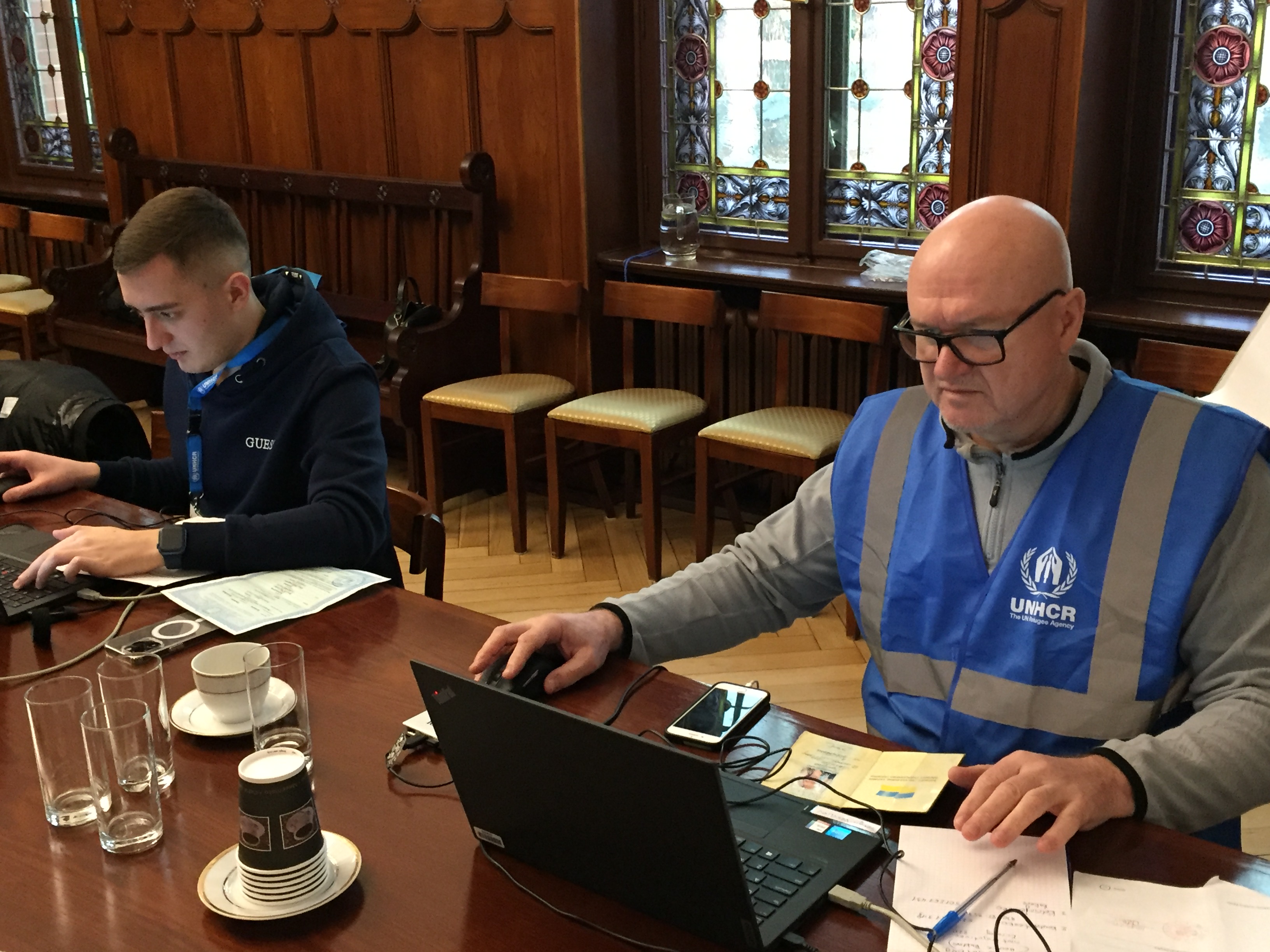 Dwóch pracowników UNHCR pracuje przy rejestracji - na stole laptopy, dokumenty, zeszyt długopis, szklanki i kubeczki