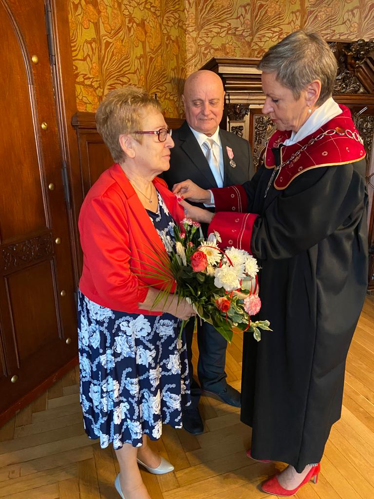 Na zdjęciu widzimy 3 osoby, Jubilatów i Prezydentkę Miasta - Prezydentka Miasta wpina medal w klapę żakietu Jubilatki trzymającej kwiaty