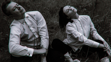 Kobieta i mężczyzna w białych koszulach siedzą na trawie i z zaduma, z zamkniętymi oczami maja głowy skierowane ku niebu.