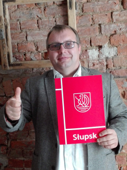 Tomasz Częścik na tle ceglanej ściany trzymający w ręce czerwoną teczkę z białym logiem Słupska i napisem Słupsk