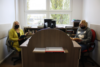 dwie kobiety siedzące przy jednym biurku, naprzeciwko siebie, w maseczkach, czytają książki, w tle okno