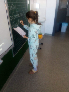 na zdjęciu widać uczennicę ubraną w piżamkę i kapcie, stojącą w klasie przy tablicy. Dziewczynka zapisuje na tablicy liczebniki: cztery, czterdzieści, czterysta.