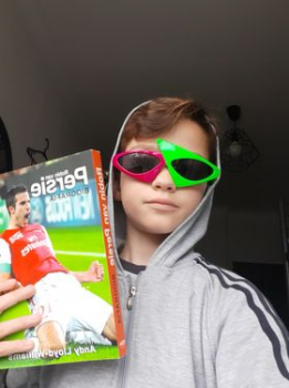 chłopiec w kolorowych okularach przeciwsłonecznych, w kapturze na głowie, trzyma w ręku książkę, na okładce piłkarz