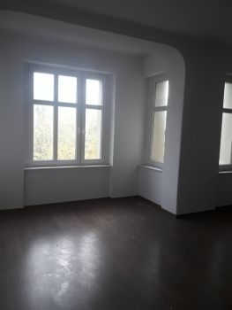 Zdjęcie przedstawia pomieszczenie mieszkalne z dużymi oknami, jasnymi ścianami i ciemną podłogą.