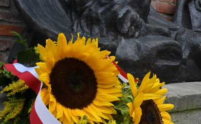 na zdjęciu widzimy fragment pomnika Powstańców Warszawskich oraz bukiet słoneczników z szarfą w kolorze barw narodowych
