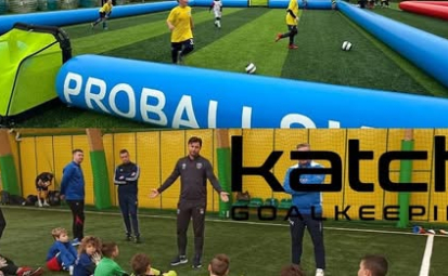 Boisko z dmuchanymi bandami i trenerzy rozmawiający z dziećmi.