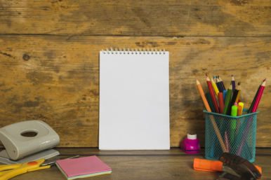 Zdjęcie przedstwia biurko z artykułami szkolnymi: dziurkacz, nożyczki, zszywacz, kredki w pojemniku i notatnik.