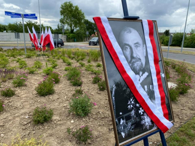 Na zdjęciu widzimy flagi Rzeczypospolitej Polskiej, tabliczki z nazwą ronda - RONDO GEN. TADEUSZA ROZWADOWSKIEGO oraz zdjęcie patrona przesłonięte szarfą w kolorach białym i czerwonym