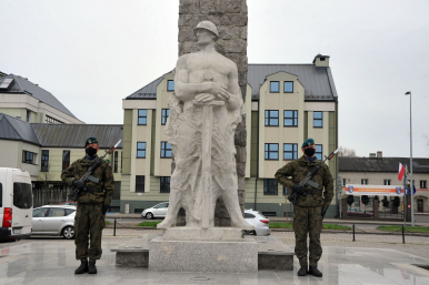 Na zdjęciu widzimy pomnik,a po jego po stronach żołnierzy w pełnym umundurowaniu z bronią. W tle kamienice, auta i flagi RP.