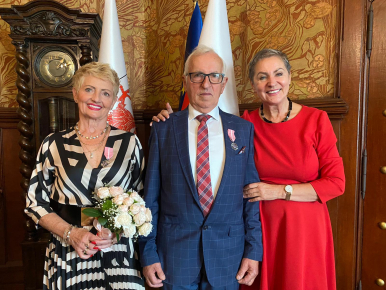 Na zdjęciu widzimy 3 osoby, Jubilatów oraz Prezydentkę. Jubilatka trzyma bukiet kwiatów, w tle zegar i flagi