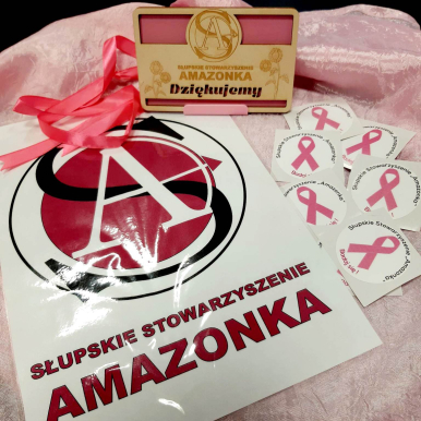 Na zdjęciu widzimy naklejki z logo AMAZONEK z napisem Słupskie Stowarzyszenie AMAZONKA oraz inne, mniejsze nalejki z napisem BADAJ SIĘ! Słupskie Stowarzyszenie "AMAZONKA". Widać też podziękowanie - Słupskie Stowarzyszenie AMAZONKA dziękujemy! (w drewnie wyryty napis i obok ozdone kwiatki)
