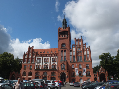 Na zdjęciu widzimy ratusz, po bokach zielone drzewa, chmury i letnie niebieskie niebo, przed ratuszem na placu zaparkowanych kilkanaście aut i przechodzących ludzi