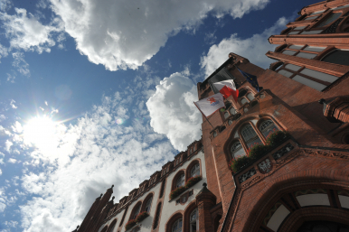 zdjęcie ratusza od dołu - widoczne niebo z chmurami oraz trzy flagi - z herbem Słupska, Polski oraz Unii Europejskiej.