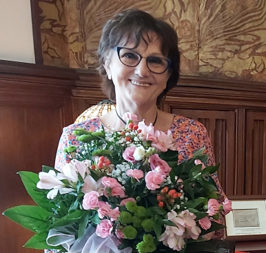 Na zdjęciu widzimy Krystynę Wrycz, wieloletnią Kierowniczkę Urzędu Stanu Cywilnego, trzymającą bukiet kwiatów, teczkę i torbę z upominkiem.