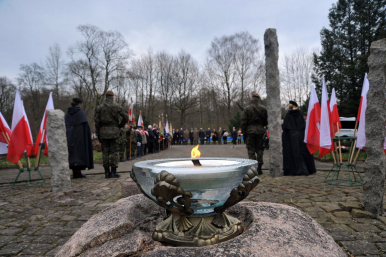 Na zdjęciu widzimy płonący okrągły znicz, w tle uczestników uroczystości miejskich: żołnierzy, osoby duchowne, harcerzy, mieszkańców miasta, władze smaorządowe, widać też flagi Rzeczypospolitej Polskiej