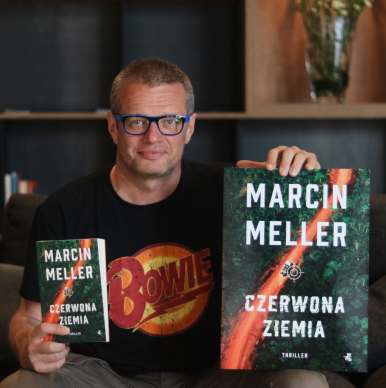 Dziennikarz Marcin Meller w pozycji siedzącej na kolanach opiera dwie książki "Czerwona Ziemia"