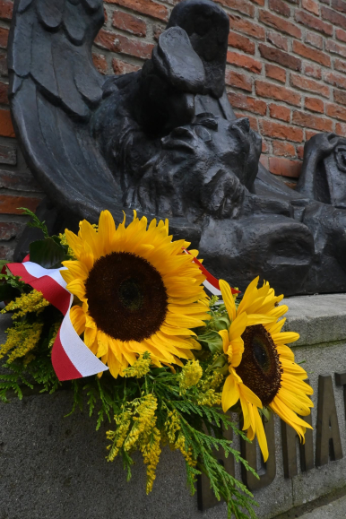 na zdjęciu widzimy fragment pomnika Powstańców Warszawskich oraz bukiet słoneczników z szarfą w kolorze barw narodowych