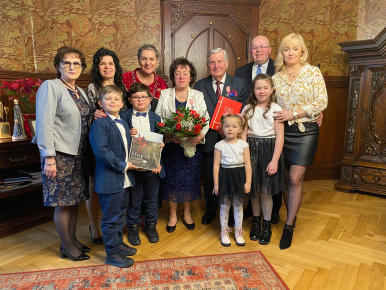 Na zdjęciu widzimy 11 osób,  w tym 4 dzieci; w tle szafka z książkami, szafa, kwiaty i ceramiczny aniołek