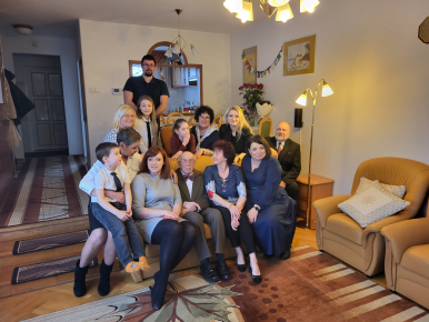 Prezydentka Miasta ,  Jubilat i kilka osób z rodziny i bliskich Pana Antoniego - siedzą na kanapie w pokoju; w tle meble, przedpokój, lustro, obrazy, na podłodze dywany, lampa.