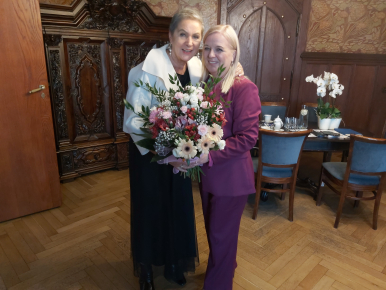 Prezydentka Słupska i Iwona Gozdek trzymające kwiaty, w tle stół z filiżankami, serwisem kawowym, szafa, meble gabinetowe