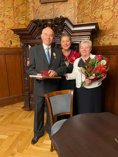 Na zdjęciu widzimy widzimy 3 osoby, 2 kobiety i 1 mężczyznę, na pierwszym planie stół i krzesło, w tle szafę; kobieta Jubilatka trzyma kwiaty, Jubilat upominki, Prezydentka Miasta stoi w środku i obejmuje Jubilatów